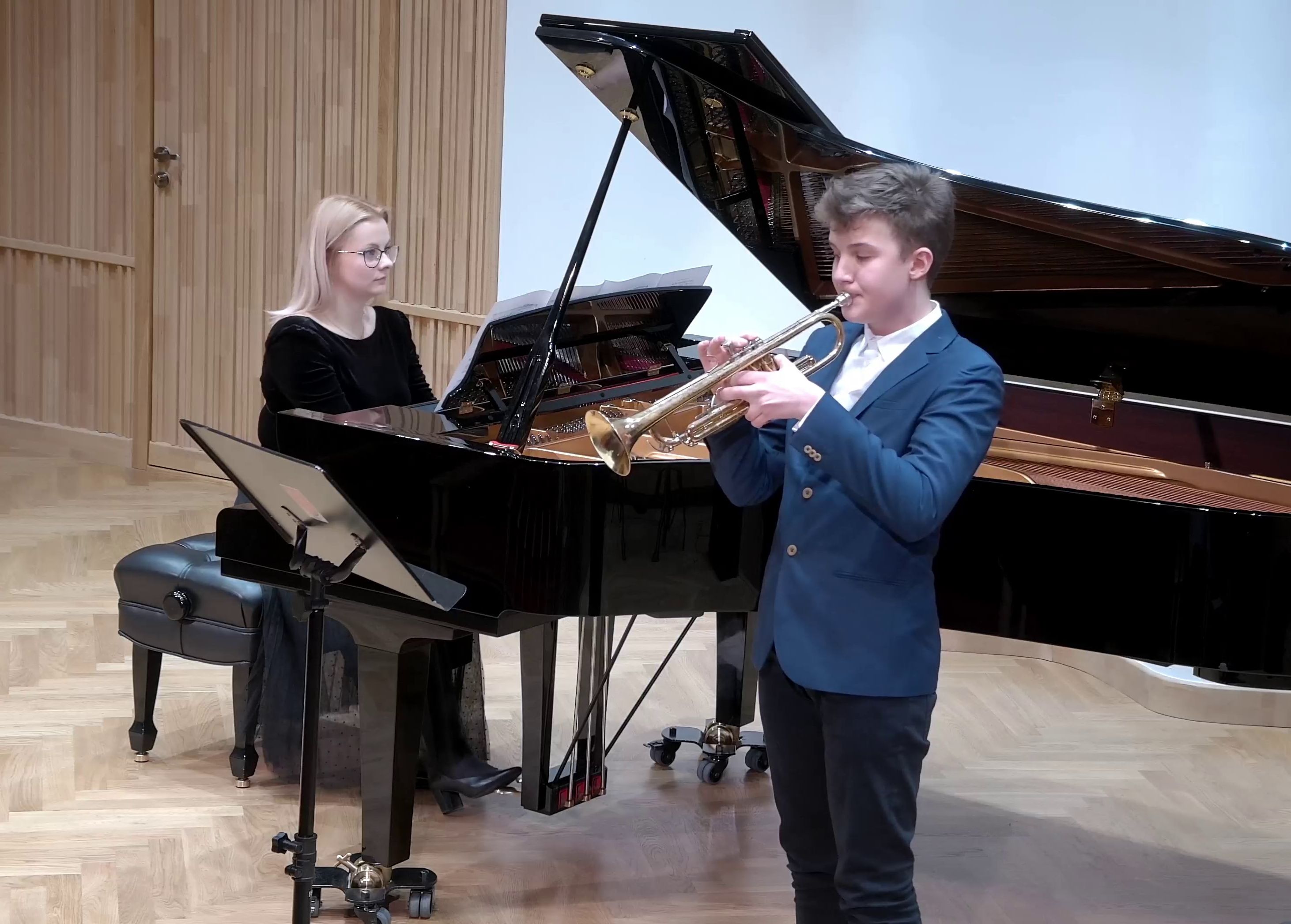 Zdjęcie. Na estradzie Sali Koncertowej występują: młody trębacz grający na trąbce oraz akompaniująca mu przy czarnym fortepianie pianistka. przed trębaczem stoi pulpit z nutami.