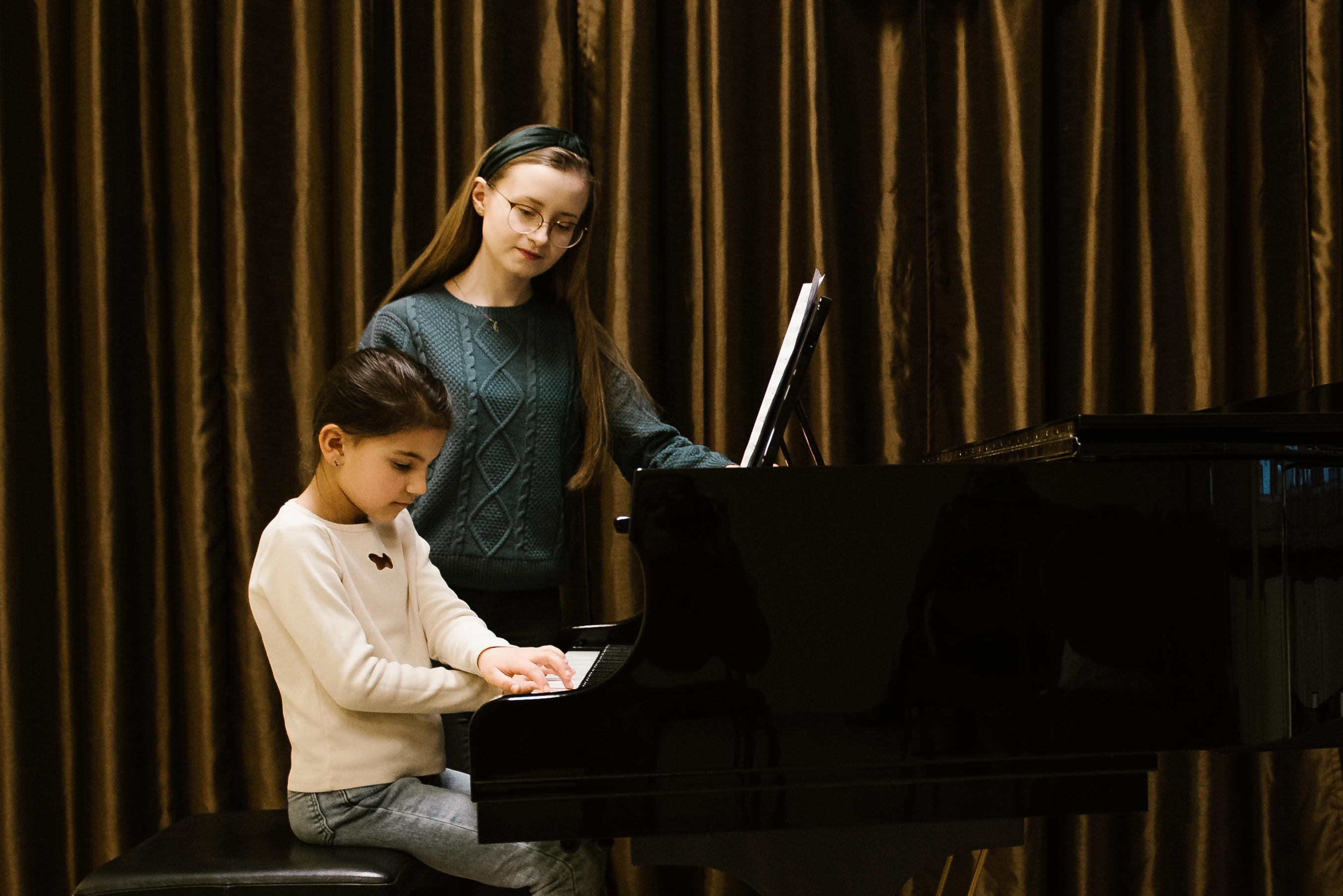 Na zdjęciu znajduje się pani Julia Rumińska-Wyrwa oraz uczennica Amelia Stefanko. Nauczycielka stoi przy czarnym fortepianie, przy którym siedzi i na którym gra uczennica. 