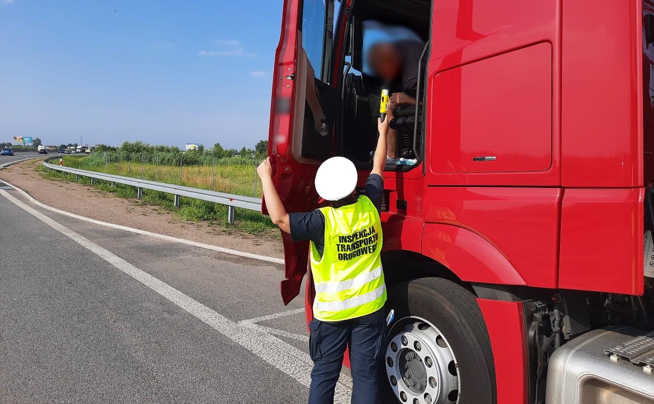 Inspektor stoi przy otwartych drzwiach kabiny ciężarówki i podstawia alkomat w kierunku twarzy kierowcy ciężarówki.