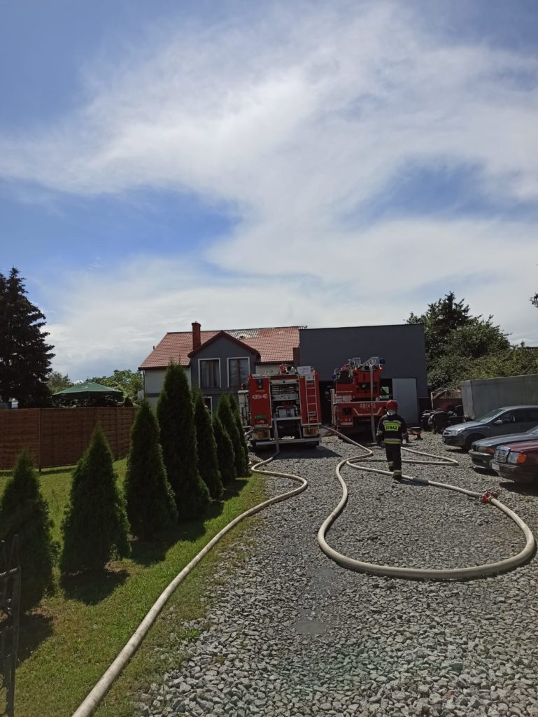 Strażak na tle samochodów pożarniczych, a w głębi palący się dom
