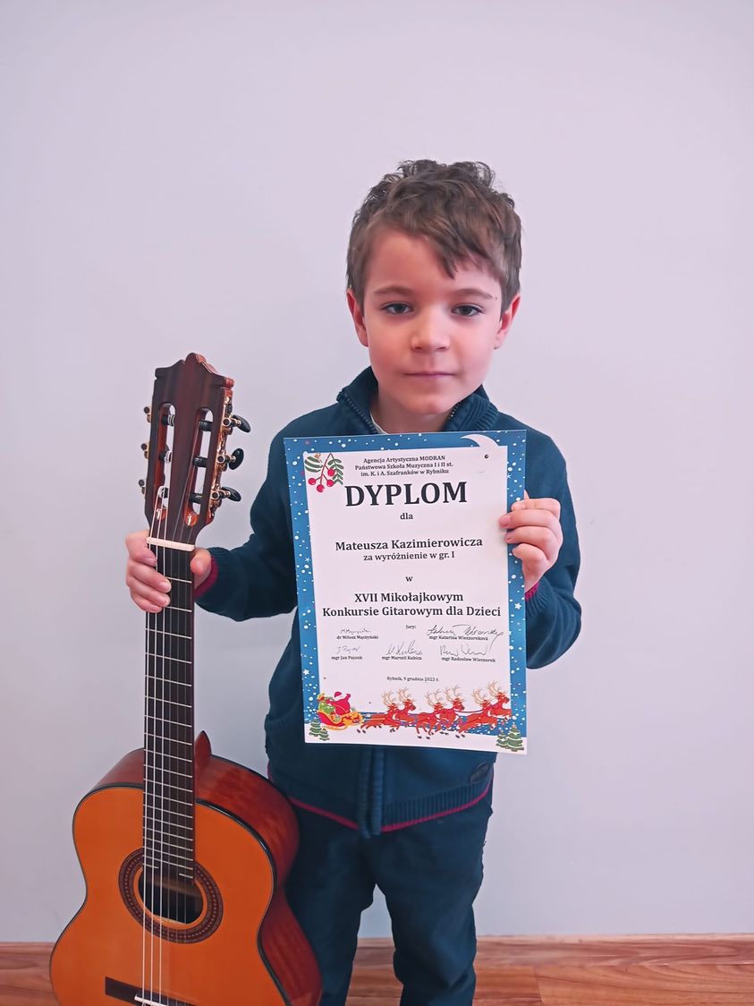 Na zdjęciu widoczny chłopczyk trzymający w lewej ręce dyplom konkursowy, w prawej gitarę. 