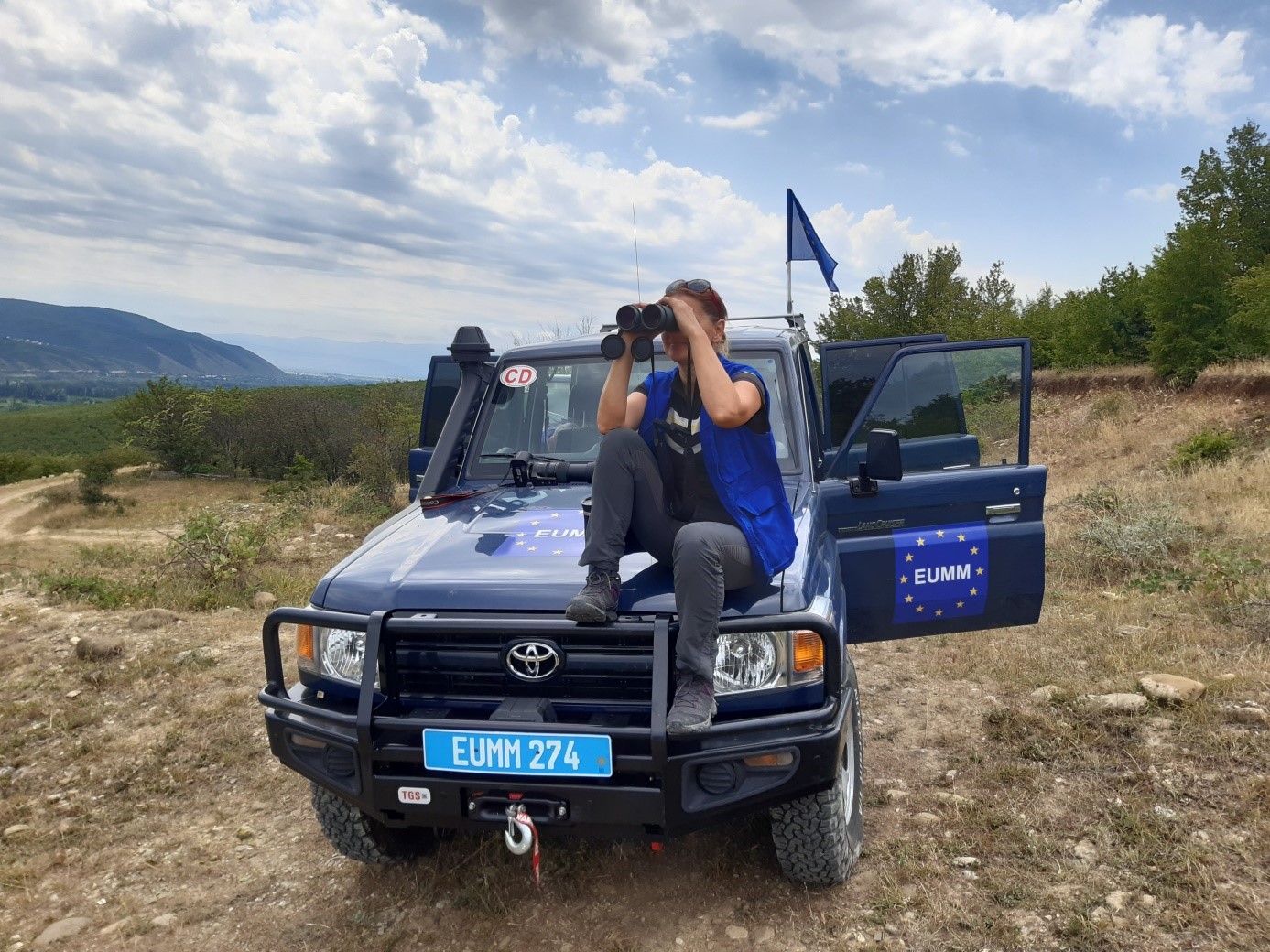 zdjęcie funkcjonariuszki, która siedzi na masce samochodu terenowego i patrzy przez lornetkę.