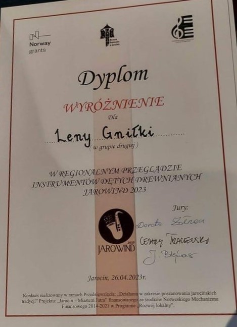 Zdjęcie ma białe tło. Nazwisko wyróżnionej uczennicy znajduje się na środku i zostało wpisane ręcznie. Poniżej widać logo przeglądu. Po prawej stronie są podpisy jury. 