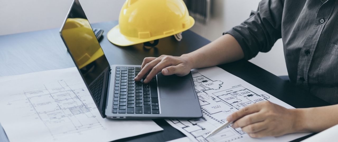 Na obrazku widać osobę siedzącą przy biurku. Osoba pracuje przy komputerze i jednocześnie trzyma długopis nad planami budynku. Na biurku leży też kask ochronny, który noszą osoby pracujące na budowie.