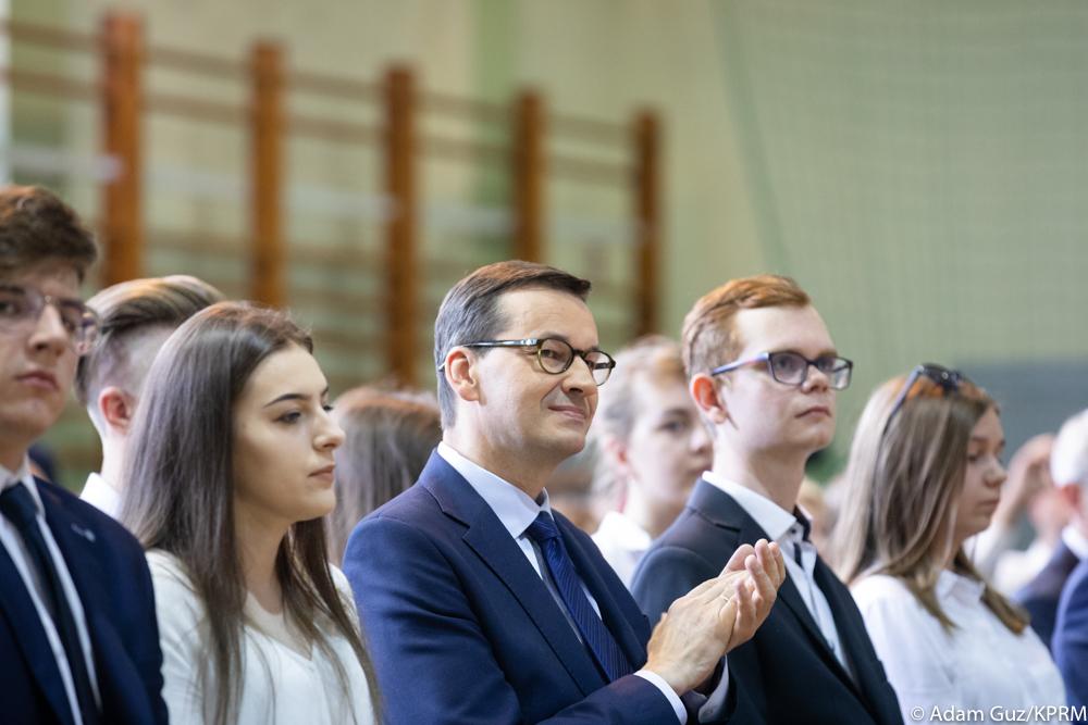 Premier siedzi wśród uczniów na zakończeniu roku szkolnego.