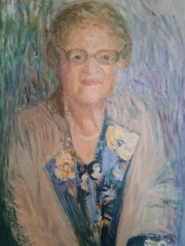 Fotografia obrazu patronki szkoły - Janiny Garści. Obraz namalowała Bernadeta Cieslewicz - Orawiec we wrześniu 1997 roku. Fotografia przedstawia około 60-letnią kompozytorkę, ubraną w jasny kostium z połami koloru granatowego w kwiaty. Na szyi ma perłowe korale. Kobieta uśmiechnięta, o jasnych krótkich włosach, w okularach. Tło wielokolorowe, pastelowe.