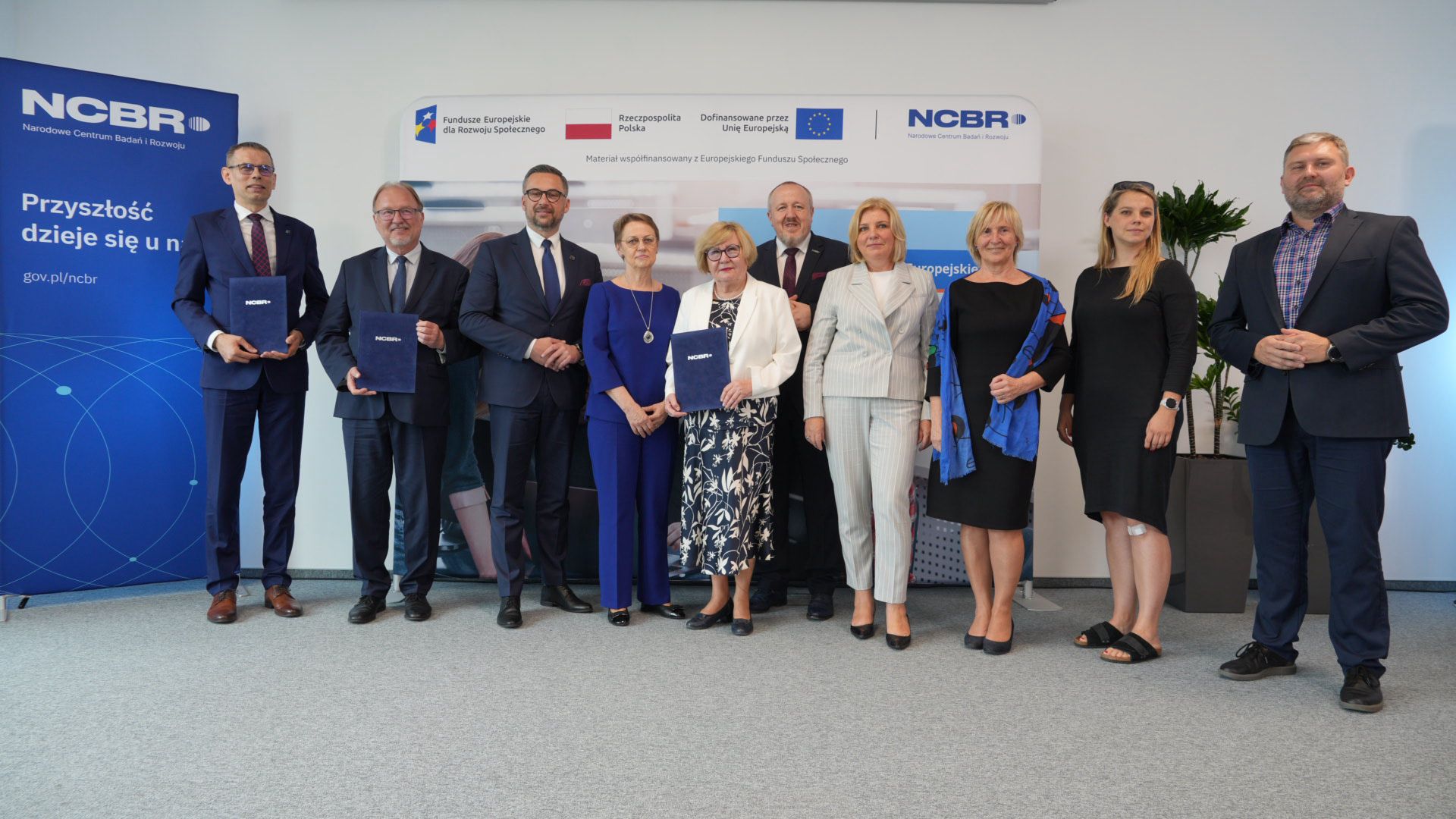 Pod ścianą stoi grupa dziesięciu osób, za nimi i z boku banery z napisem NCBR.