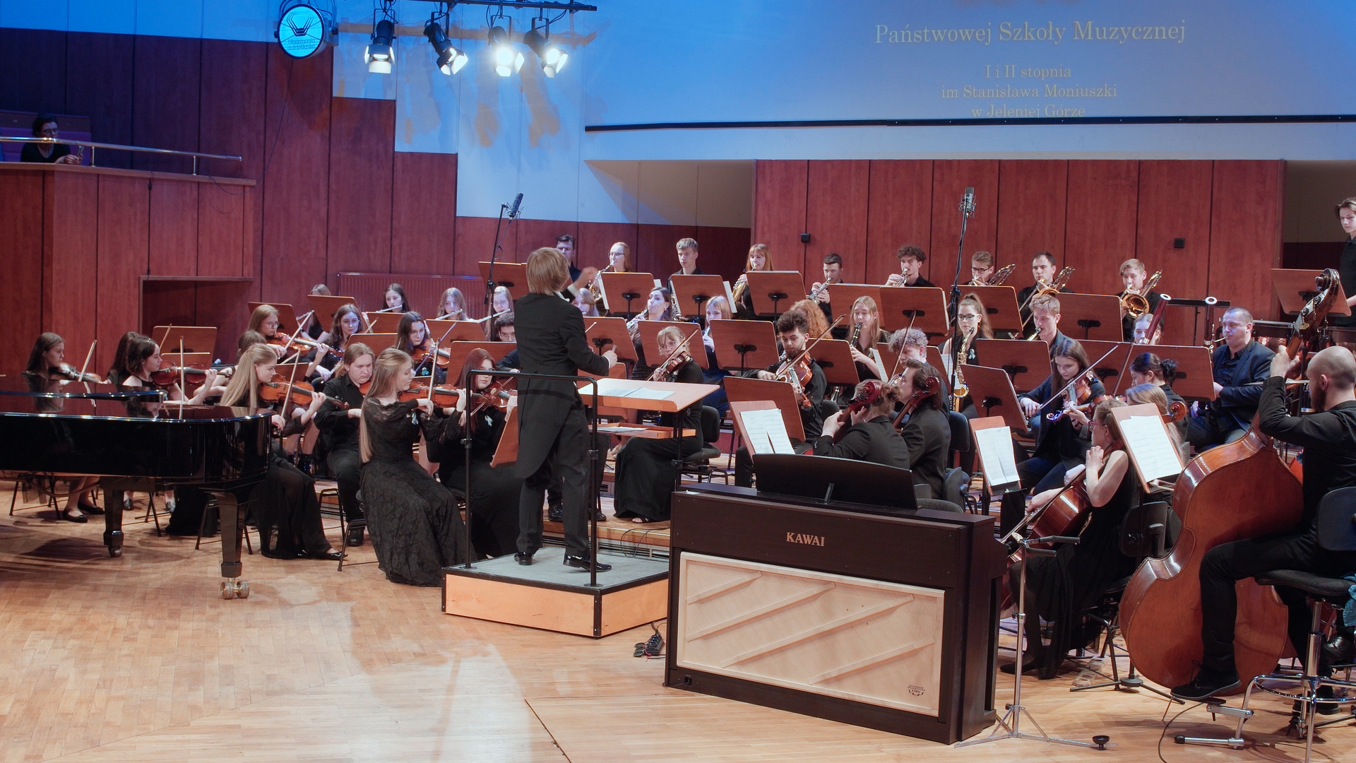 Zdjęcie zrobione podczas występu orkiestry symfonicznej PSM II stopnia na 75 leciu szkoły muzycznej w sali filharmonii Dolnośląskiej. Na zdjęciu dyrygent oraz członkowie orkiestry grający na instrumentach.