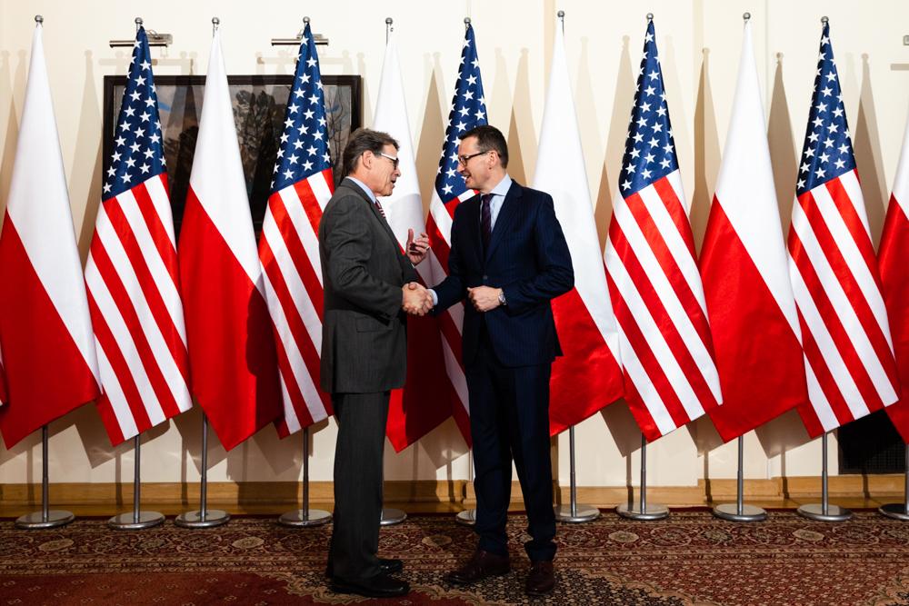 Premier Mateusz Morawiecki i sekretarz Rick Perry ściskają sobie dłonie na tle flag polskich i amerykańskich. 