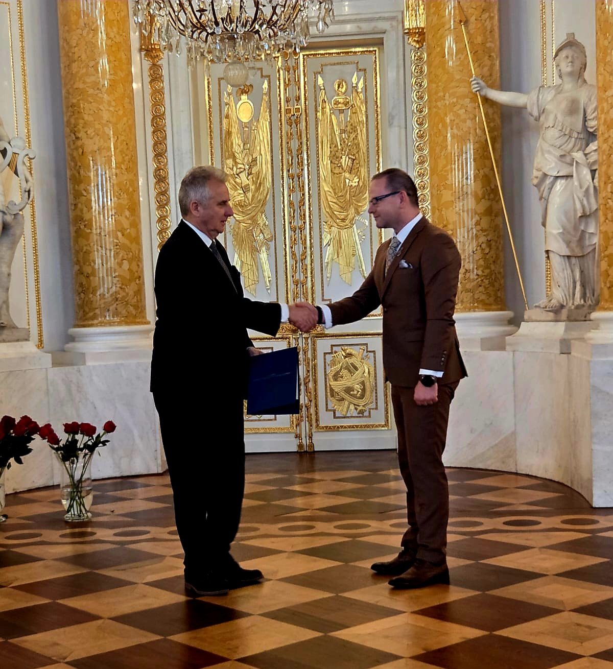 Pełne ujęcie przedstawia dwóch mężczyzn stojących i ściskających sobie dłonie w formalnym, bogato zdobionym wnętrzu.