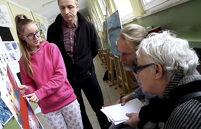 Na zdjęciu widać trzech nauczycieli z Państwowego Liceum Sztuk Plastycznych w Zielonej Górze, którzy ogladają na szkolnym korytarzu pracę malarską trzymaną w rękach przez uczennicę.