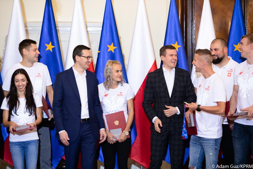 Uśmiechnięci premier Mateusz Morawiecki oraz minister Witold Bańka stoją wśród śmiejących się medalistów.