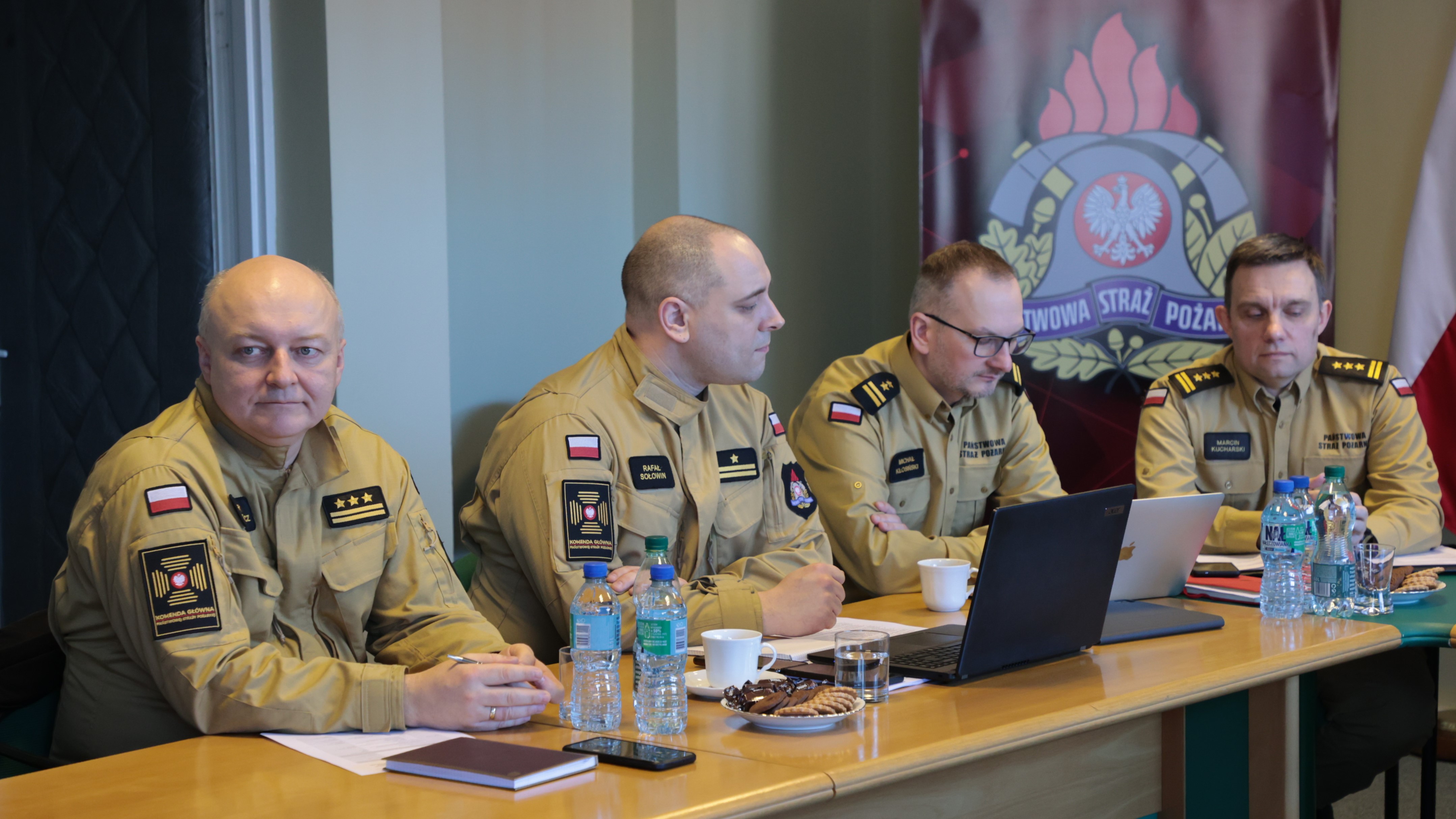 Czterech funkcjonariuszy KG PSP siedzi przy stole podczas spotkania, za nimi stoi baner z logo PSP.