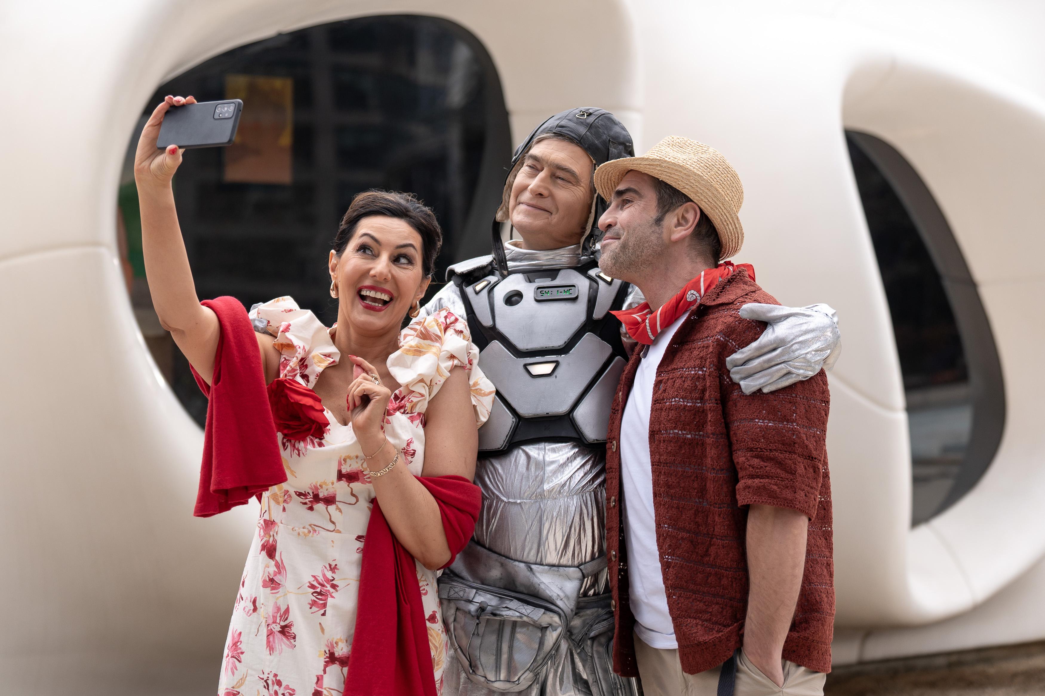 Uśmiechnięty mężczyzna w stroju astronauty stoi między mężczyzną a kobietą, razem pozują do zdjęcia, kobieta trzyma telefon kmórkowy