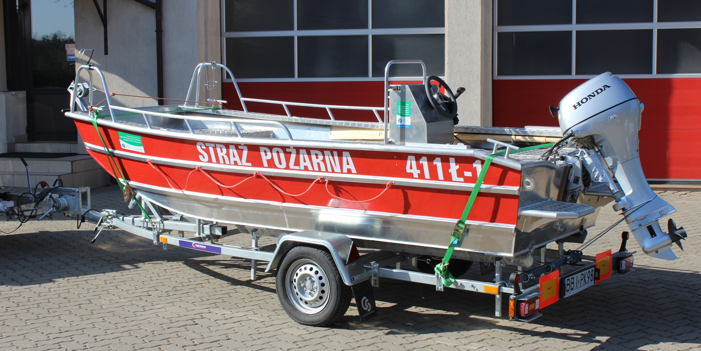 Zdjęcie przedstawia pożarniczą łódź śrubowa płaskodenna Marims 400 stojącą na przyczepie służącej do jej transportu na tle strażnicy.