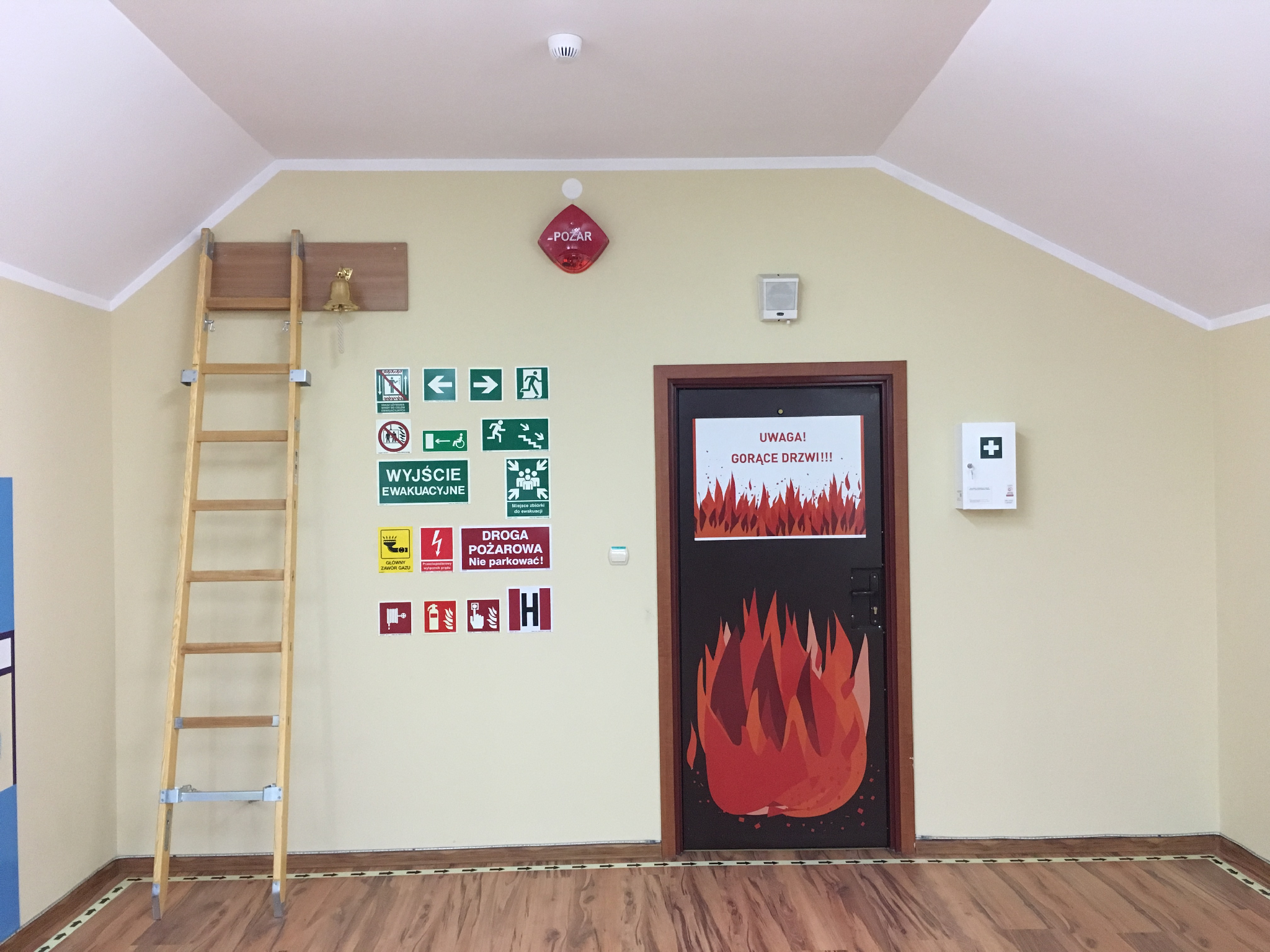 Zdjęcie przedstawia salę edukacyjną, po prawej stronie ciepłe drzwi symulujące pożar za ścianą, po lewej drabina a nad nią dzwonek do sygnalizacji alarmu