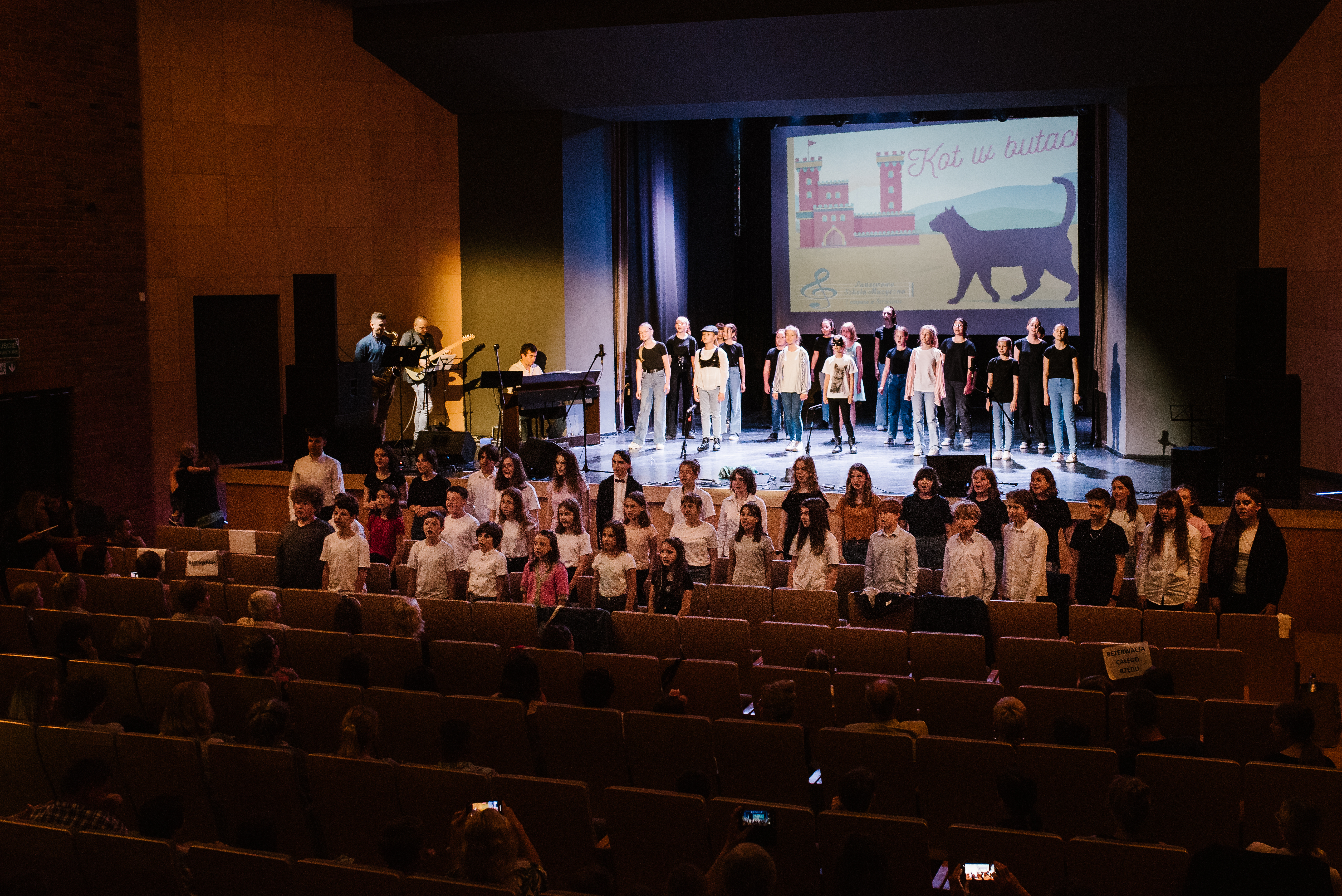 Na zdjęciu widoczna jest scena oraz kilka rzędów krzeseł. Zarówno na scenie jak i w trzech pierwszych rzędach stoją śpiewający uczniowie. Po lewej stronie widoczny jest grający zespół muzyczny.
