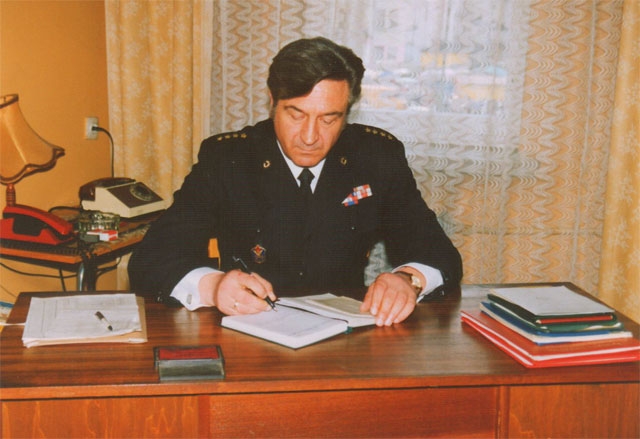 Antoni Bienias siedzi w mundurze ze stopniem starszego kapitana przy biurku i pisze w zeszycie. Na biurku leżą dokumenty.