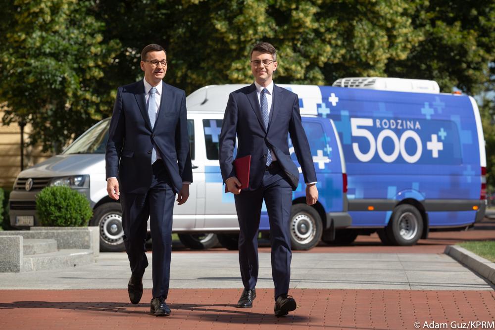 Premier Mateusz Morawiecki idzie z ministerm Piotrem Müller, a w tle Bus 500++.