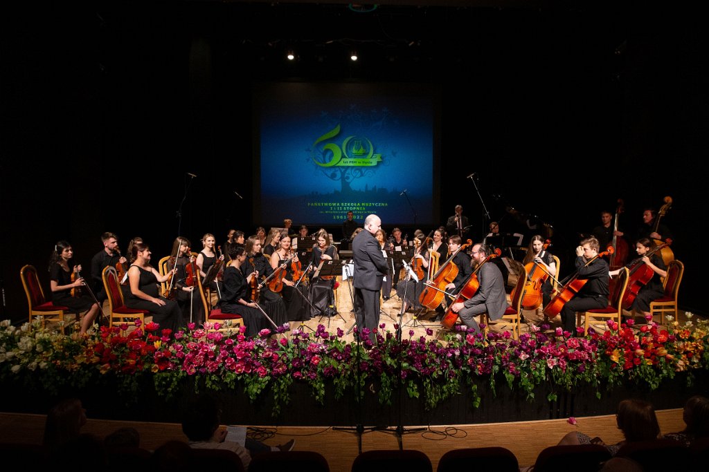 Zdjęcie: Jubileusz 60-lecia PSM w Nysie - część oficjalna - 13 maja 2022 - Nyski Dom Kultury - scena - Orkiestra Kameralna pod dyrekcją Krzysztofa Sali. W skład orkiestry wchodzą uczniowie, nauczyciele i absolwenci szkoły. W tle wyświetlone na wiszącym ekranie logo jubileuszu. Na całym obrzeżu sceny bukiety różnokolorowych kwiatów. Kliknięcie na zdjęcie przeniesie do prezentacji cyklu zdjęć z koncertu.