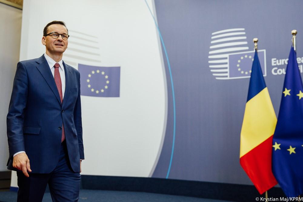 Premier Mateusz Morawiecki idzie, a za nim ścianka wizyjna i flagi.
