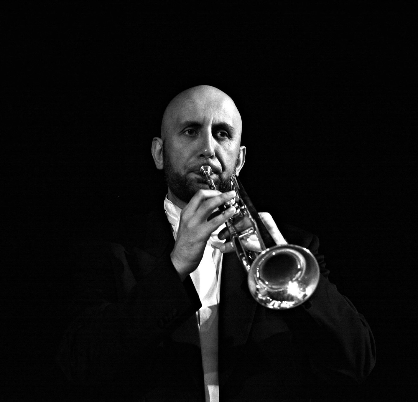 Postać trębacza na czarnym tle, ubranego w strój koncertowy, czarny frak, biała koszula, biała muszka, zwrócony frontem do widza, grający na srebrnej trąbce.