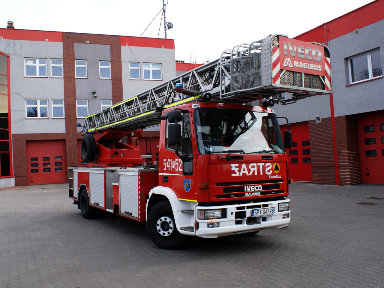 Zdjęcie przedstawia samochód specjalny drabinę mechaniczną marki Iveco Magirus w kolorze czerwonym na garażu komendy. Z tyłu przyczepa z czerwoną plandeką.