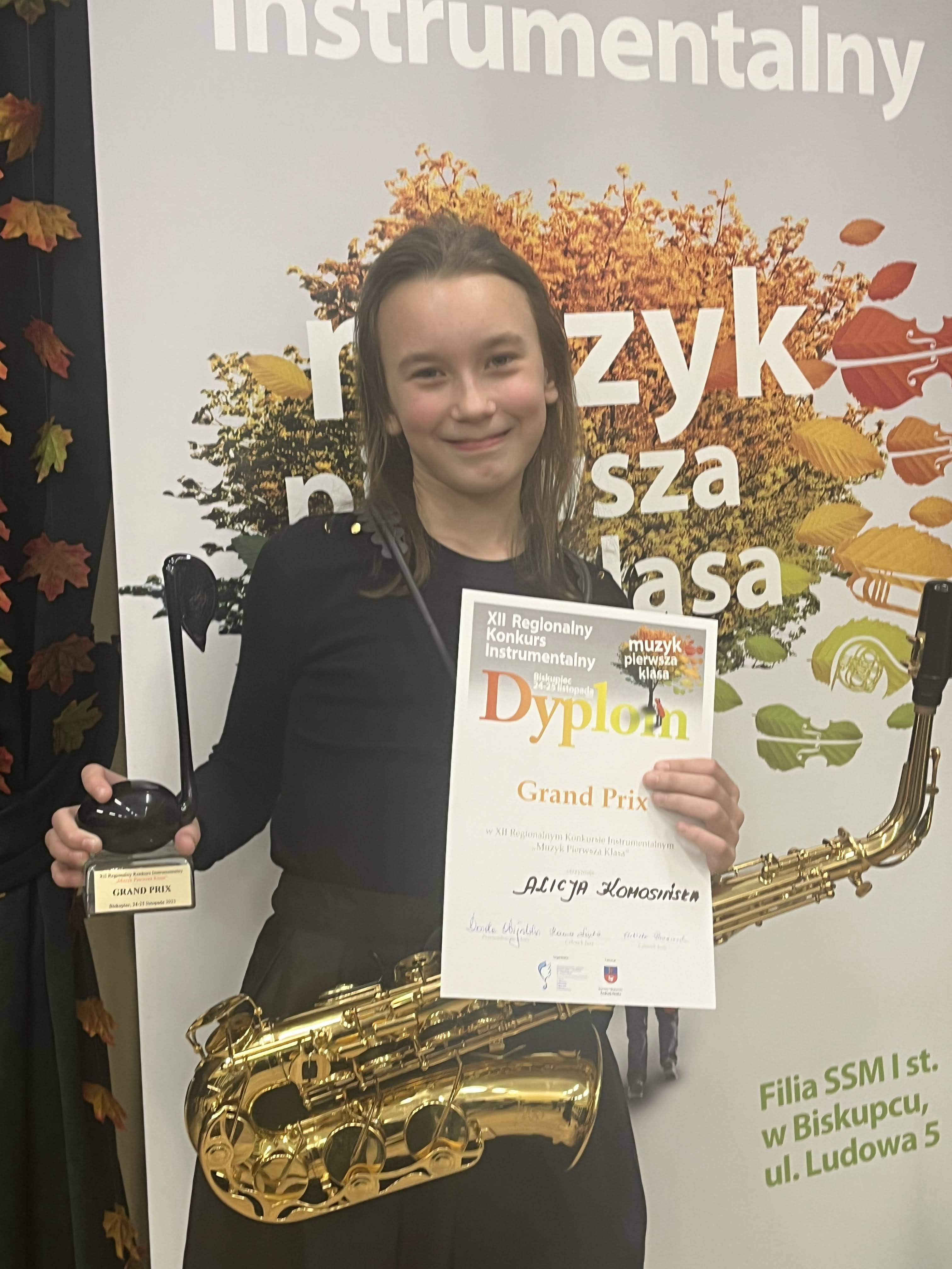 Na zdjęciu widać dziewczynkę ze złotym saksofonem ubraną w czarną sukienkę. Dziewczynka trzyma w ręku dyplom oraz statuetkę przedstawiającą czarną nutę. W tle widać baner z napisem XII Regionalny Konkurs Instrumentalny "Muzyk pierwsza klasa"