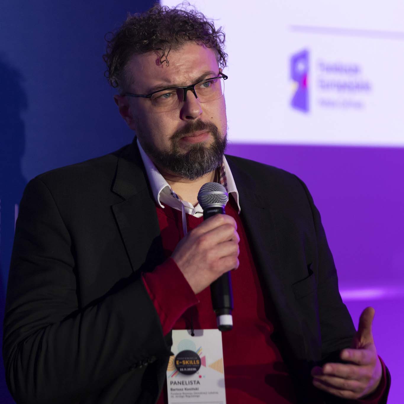 Pan Bartosz Kosiński trzyma w ręku mikrofon i przemawia.