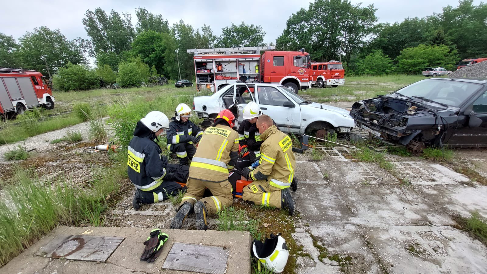 Grupa strażaków podczas ćwiczeń na otwartym terenie, w tle rozbite samochody a za nimi 3 samochody ratowniczo - gaśnicze 