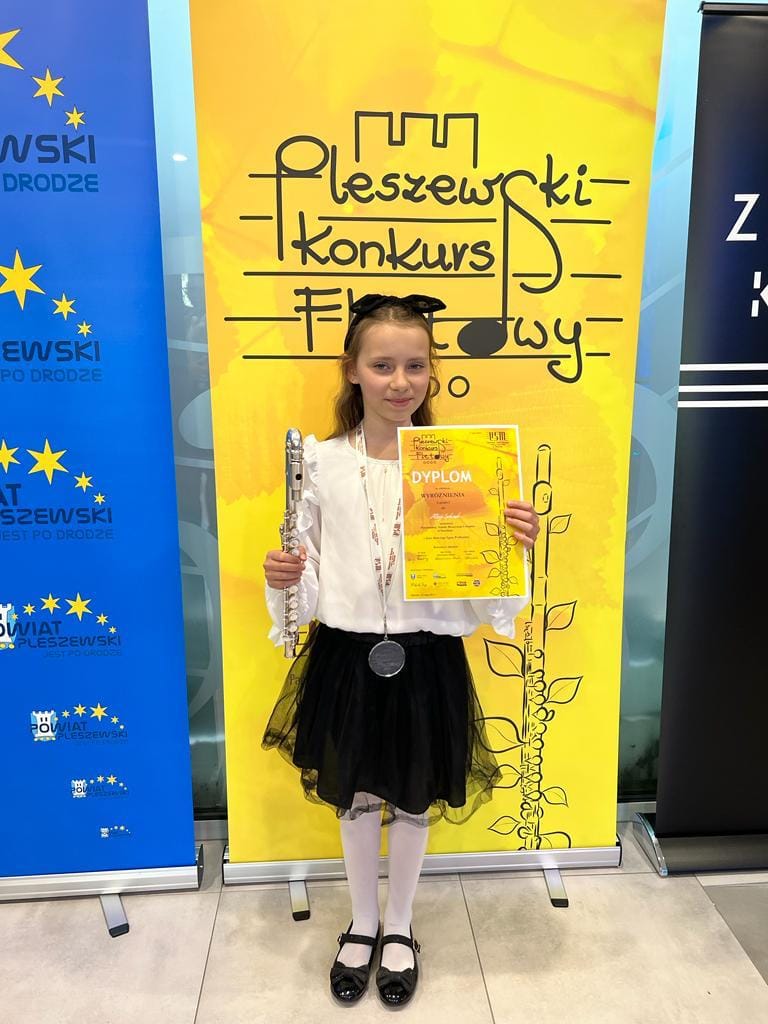 Na zdjęciu widoczna jest uśmiechnięta i elegancko ubrana uczennica, trzymająca w rękach flet oraz dyplom konkursowy. Stoi na tle żółtego baneru konkursowego.