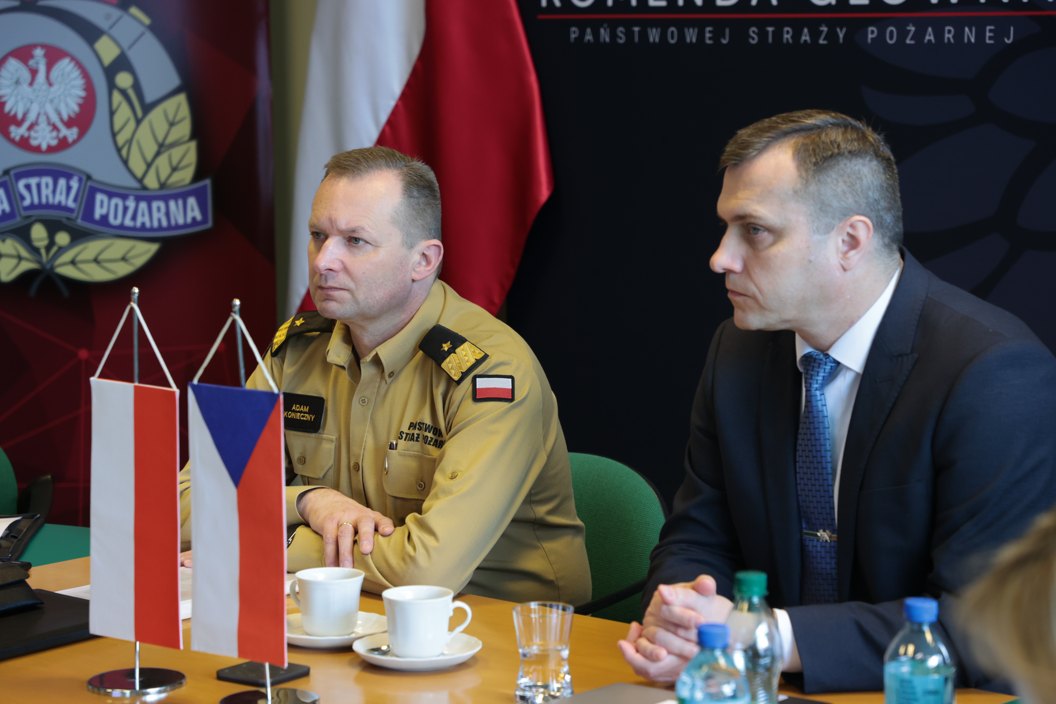 Zastępca komendanta głównego PSP wraz z przedstawicielem Czech siedzą przy stole podczas spotkania grupy roboczej, przed nimi stoją flagi Polski i Czech