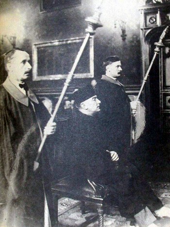 Czarno-białe zdjęcie z ceremonii przyznania Honoris Causa w sali UJ. Postać główna: mężczyzna u stroju profesora uniwersytetu siedzi na zdobionym krześle. Po jego bokach dwóch mężczyzn w togach trzymających insygnia w postaci długich lasek.