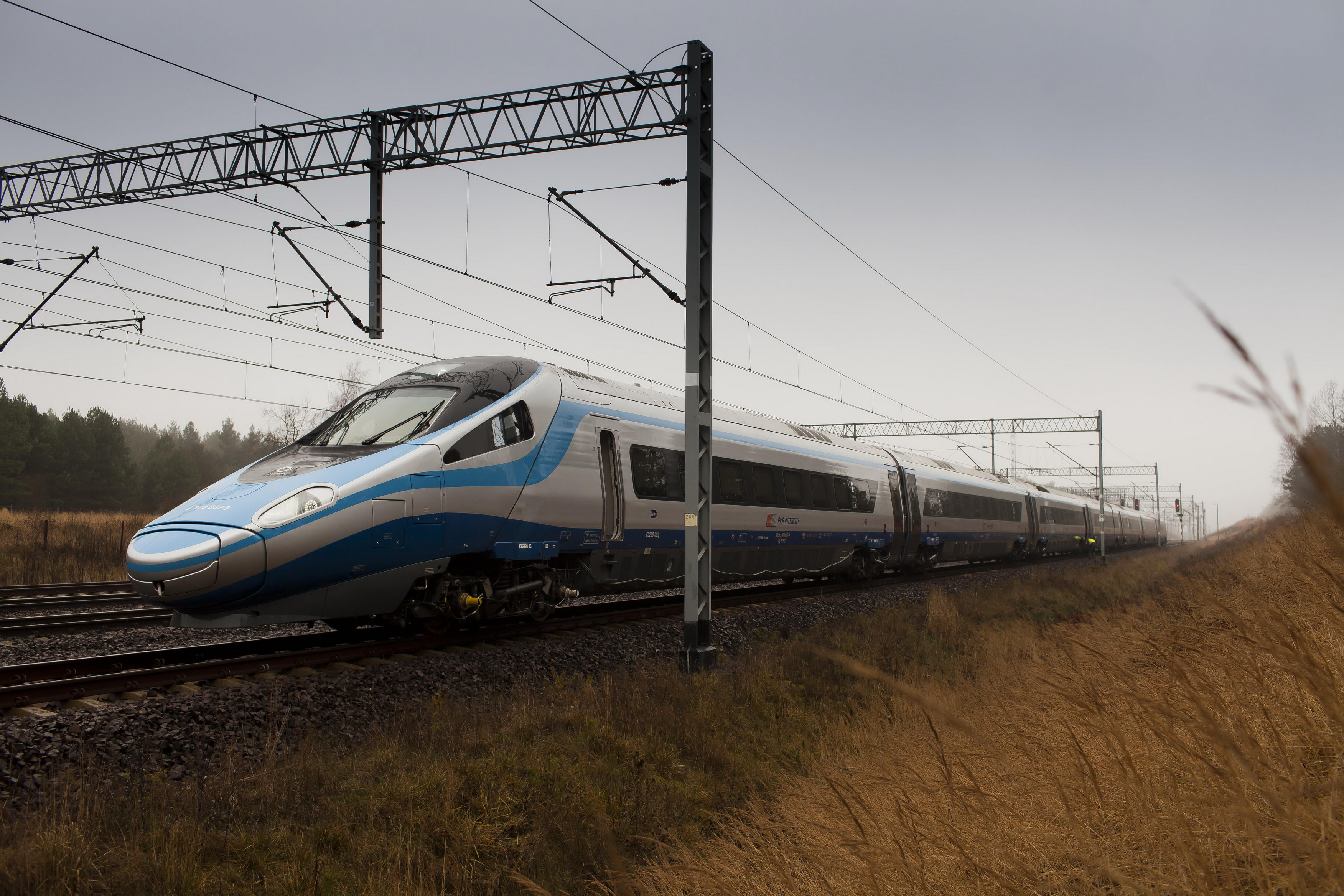 Pociąg PKP Intercity. Na pierwszym planie pociąg Pendolino jadący po torach przez pola. W tle widać słupy sieci trakcyjnej.