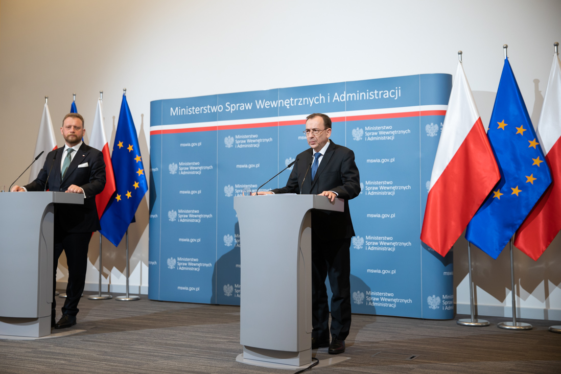 Na zdjęciu widać stojących ministrów Mariusza Kamińskiego oraz ministra Łukasza Szumowskiego podczas konferencji prasowej w MSWiA. W tle flagi Polski i UE.