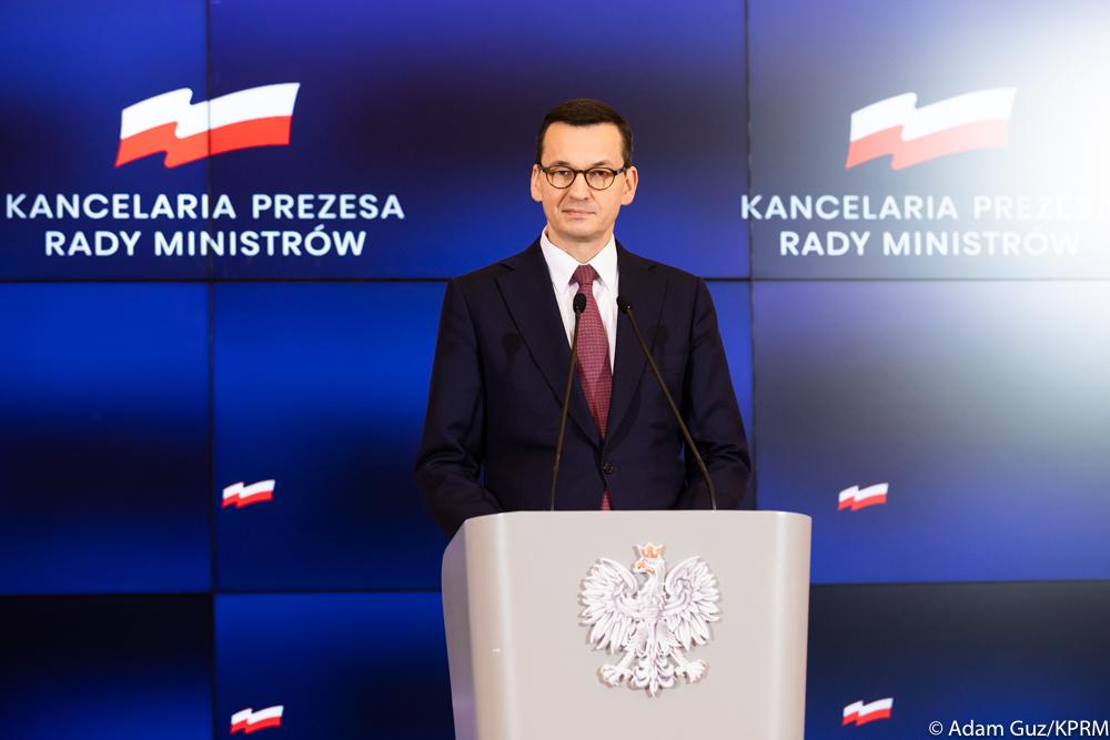 Premier Mateusz Morawiecki przemawia na mównicy na tle ścianki wizyjnej z logiem KPRM.
