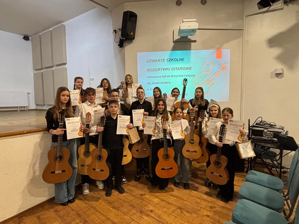 Zdjęcie przedstawia wszystkich uczestników szkolnych rozgrywek. Uczniowie stoją przed sceną i trzymają gitary oraz dyplomy. 