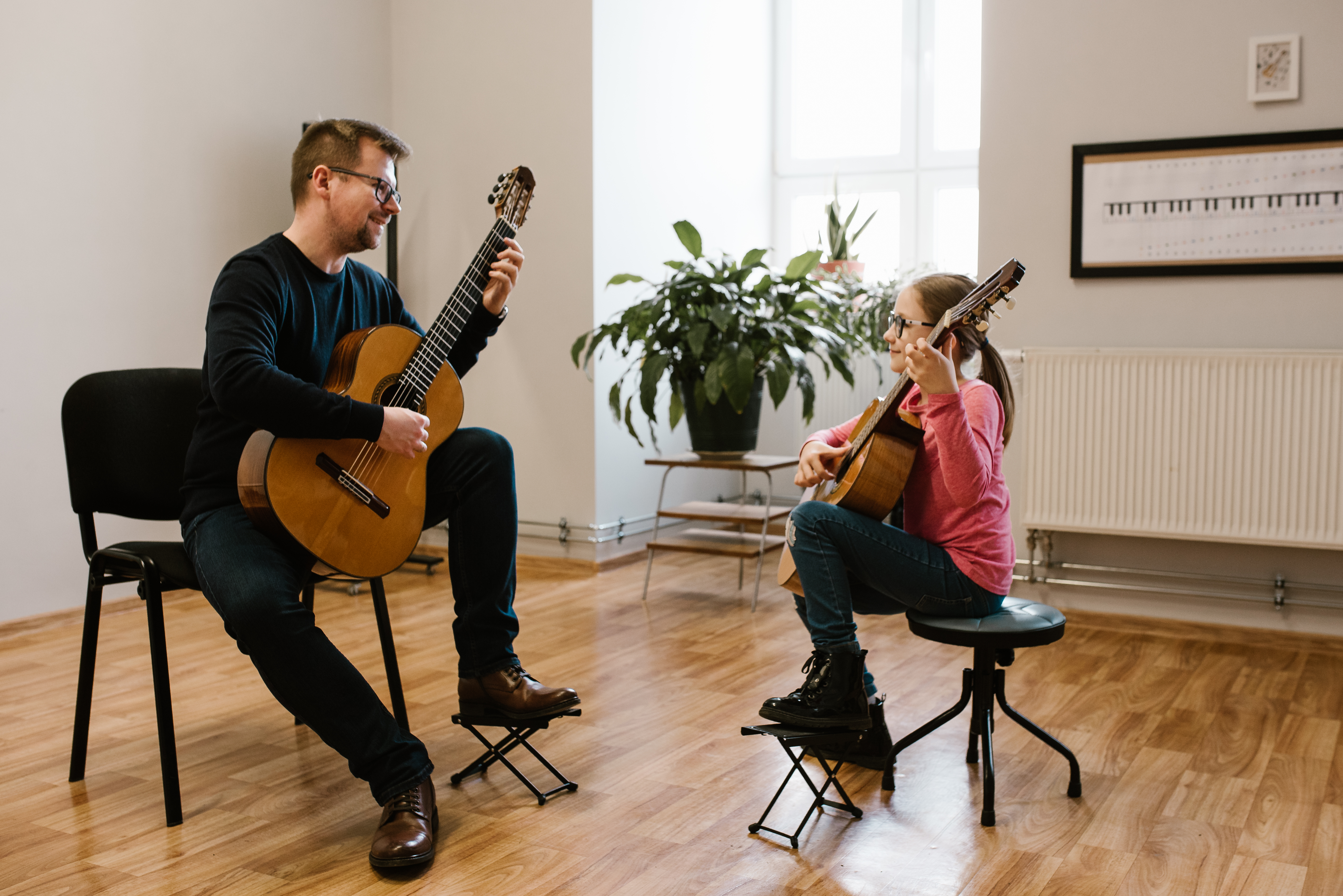Na zdjęciu znajduje się nauczyciel Łukasz FIgiel oraz uczennica Oliwia Kasprzyszak grający na gitarach. W tle znajduje się zielona roślina, na ścianie wisi meloplast w czarnej ramce. 
