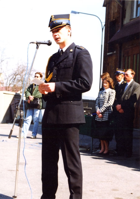 Na palcu Komendy przy ulicy Grybowskiej w Nowym Sączu stoi przed mikrofonem kpt. Maciej Halota – Dowódca JRG Nowy Sącz. W tle widoczne osoby.
