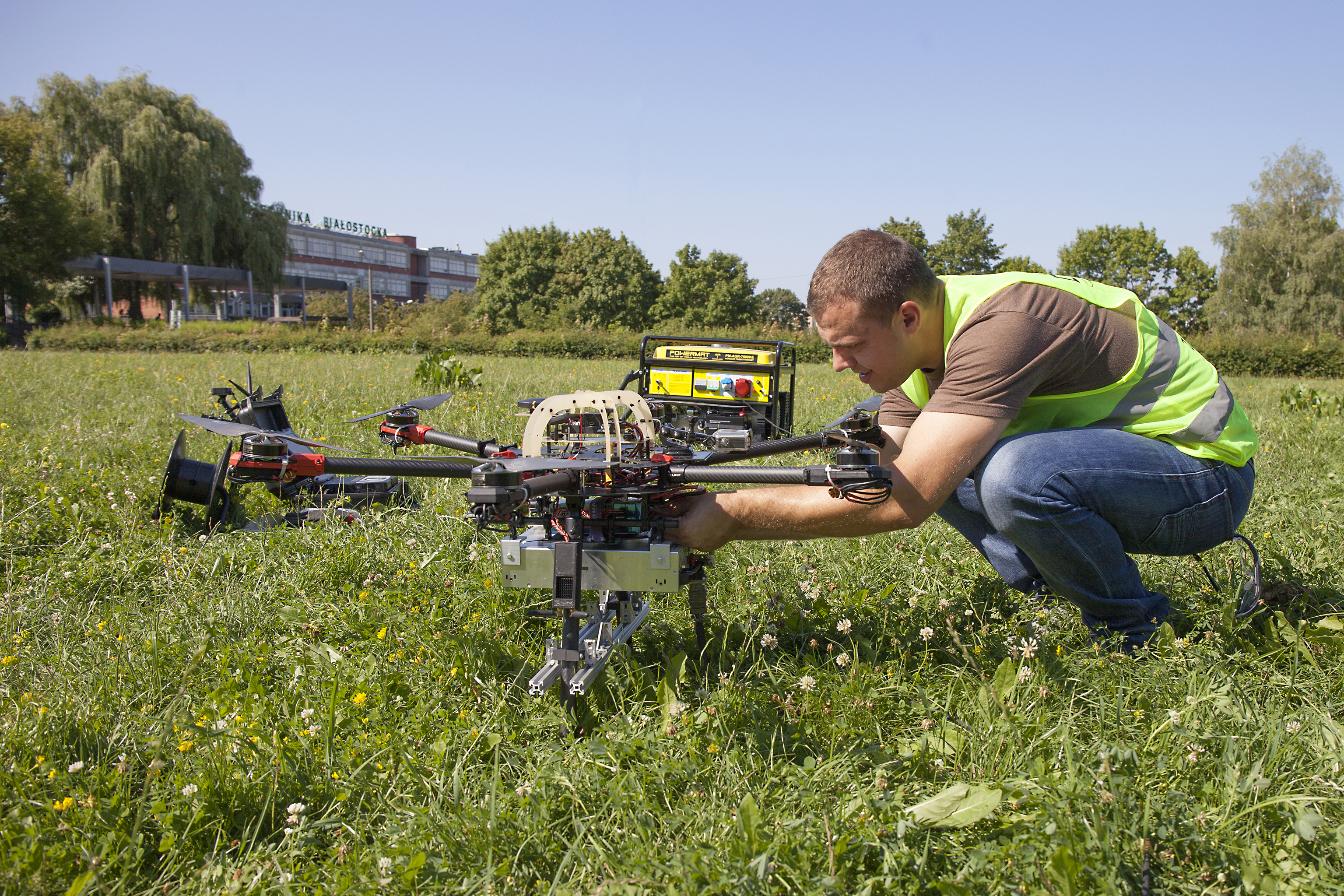 Testy elementów systemu AVAL przeprowadzone na Politechnice Białostockiej. Na zdjęciu mężczyzna w żółtej kamizelce ochronnej pracuje przy dronie na łące 