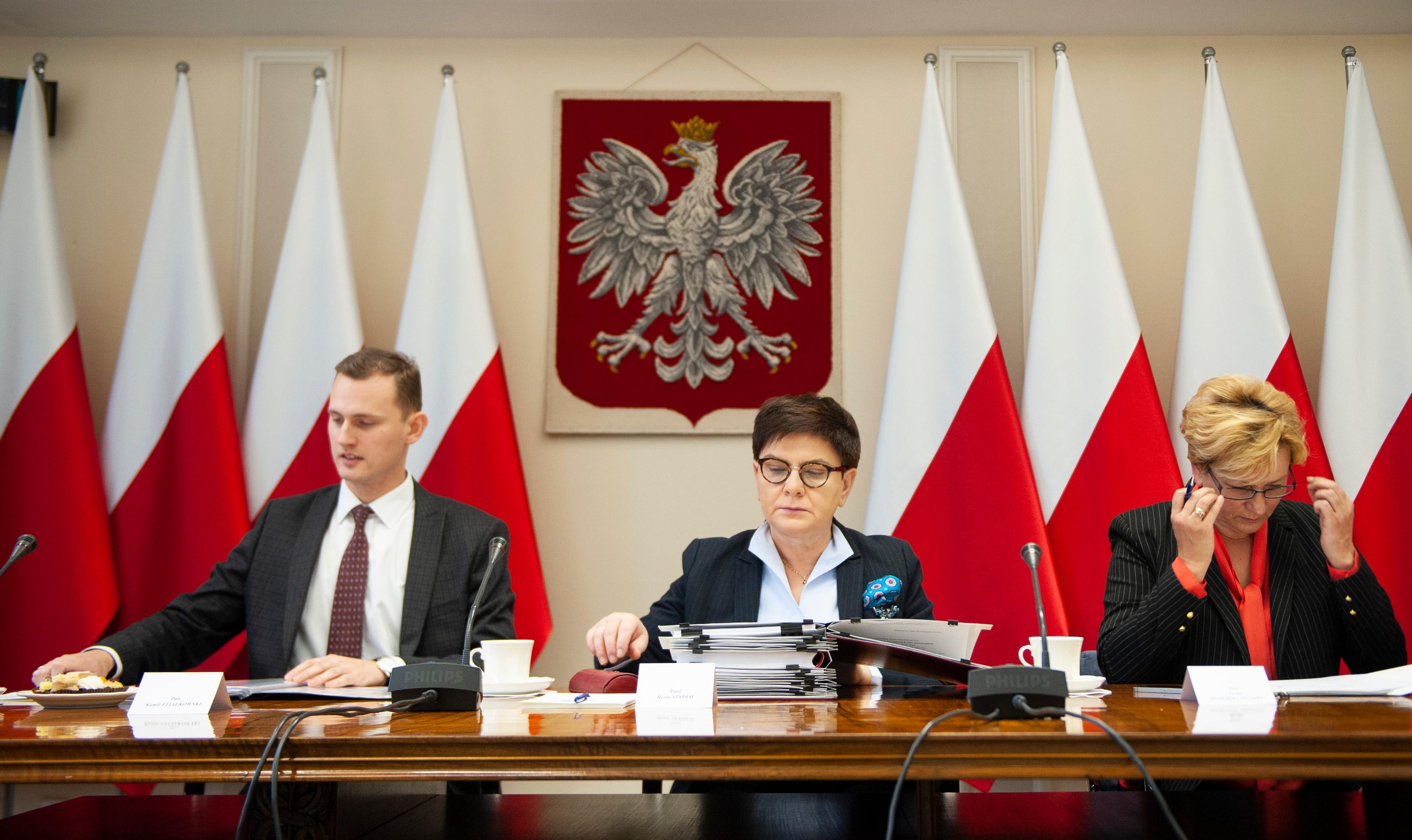 Wicepremier Beata Szydło ze współpracownikami siedzi na tle godła i biało-czerwonych flag.