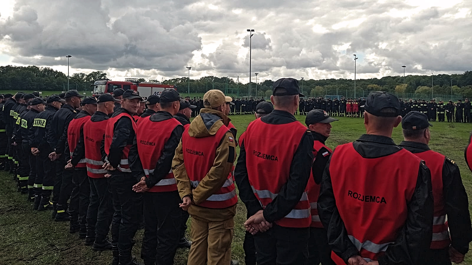 Uczestnicy ćwiczeń stojący na zbiórce podsumowującej manewry. Na pierwszym planie strażacy stojący w dwuszeregu w kamizelkach z napisem "Rozjemca".
