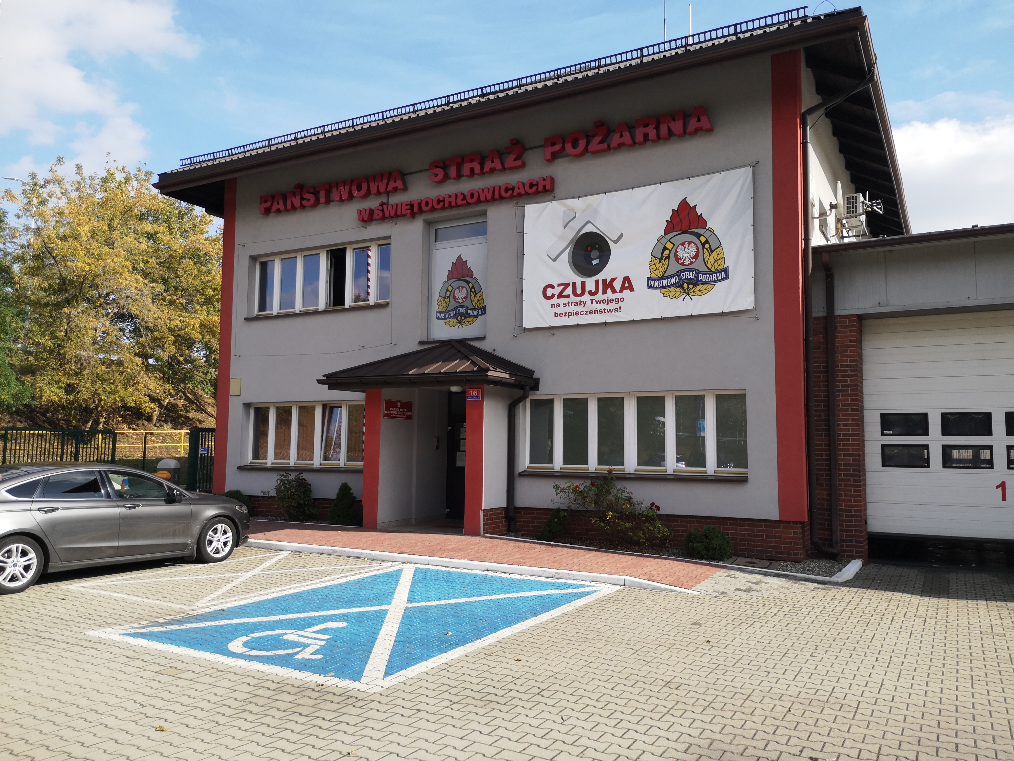 Budynek komendy od strony wejścia głównego, od frontu budynku komendy. Budynek ma parter i piętro. Jest w kolorze szarym z pionowymi pasami w kolorze czerwonym. Przed budynkiem znajduje się parking. Jedno miejsce parkingowe jest pomalowane na niebiesko ze znakiem przeznaczonym dla osób ze szczelnymi potrzebami. Obok niego są inne miejsca parkingowe. Na jednym z nich stoi samochód osobowy w kolorze szarym. Pośrodku bryły budynku na parterze znajduje się wysunięte i zadaszone wejście do budynku. Po prawej strony od bryły budynku zaczynają się garaże komendy. Połowę pierwszej bramy garażowej widać na zdjęciu.