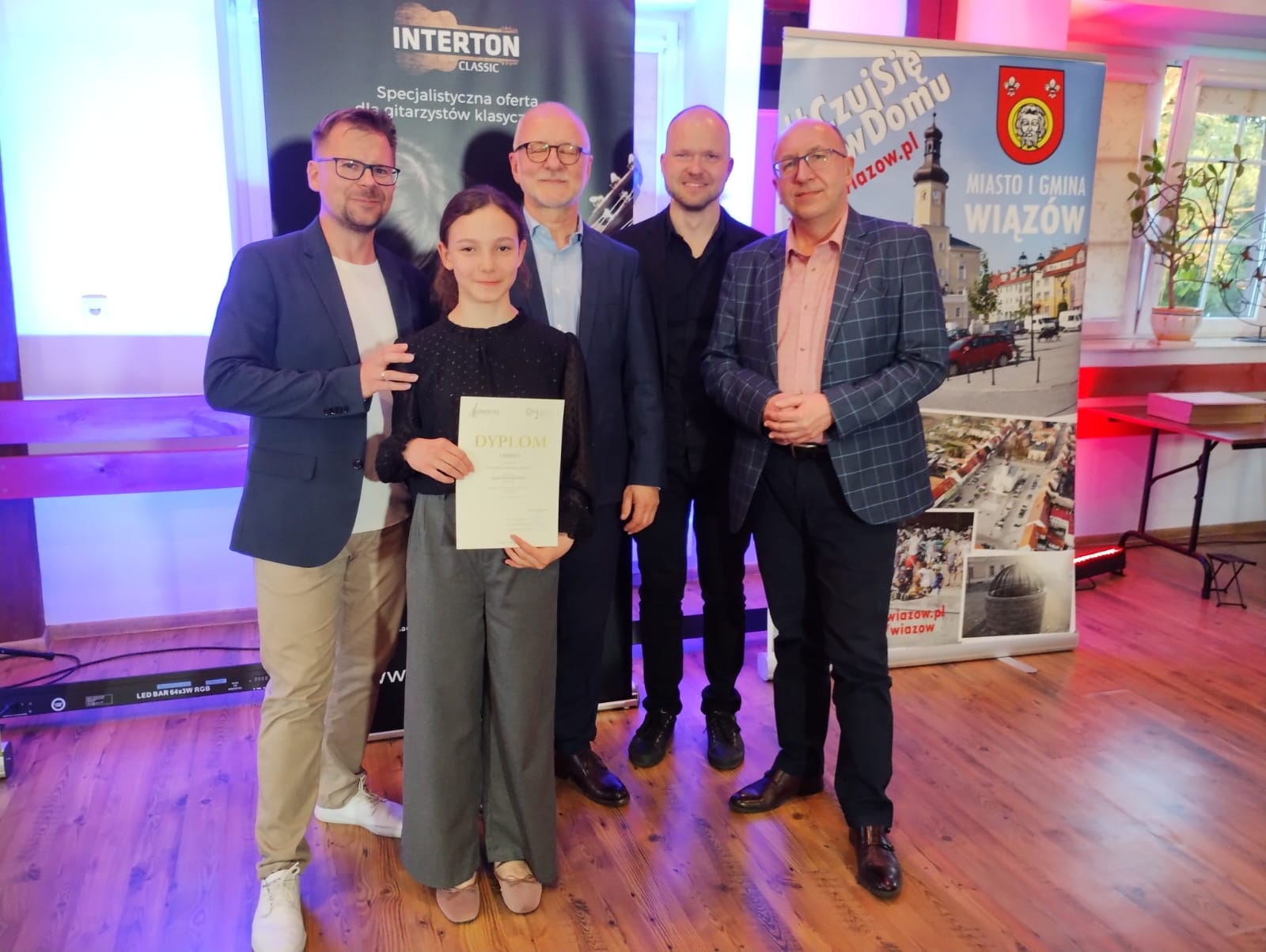 Na zdjęciu widoczna uśmiechnięta dziewczynka trzymający dyplom konkursowy w towarzystwie nauczyciela oraz trzech Panów z komisji konkursowej. 