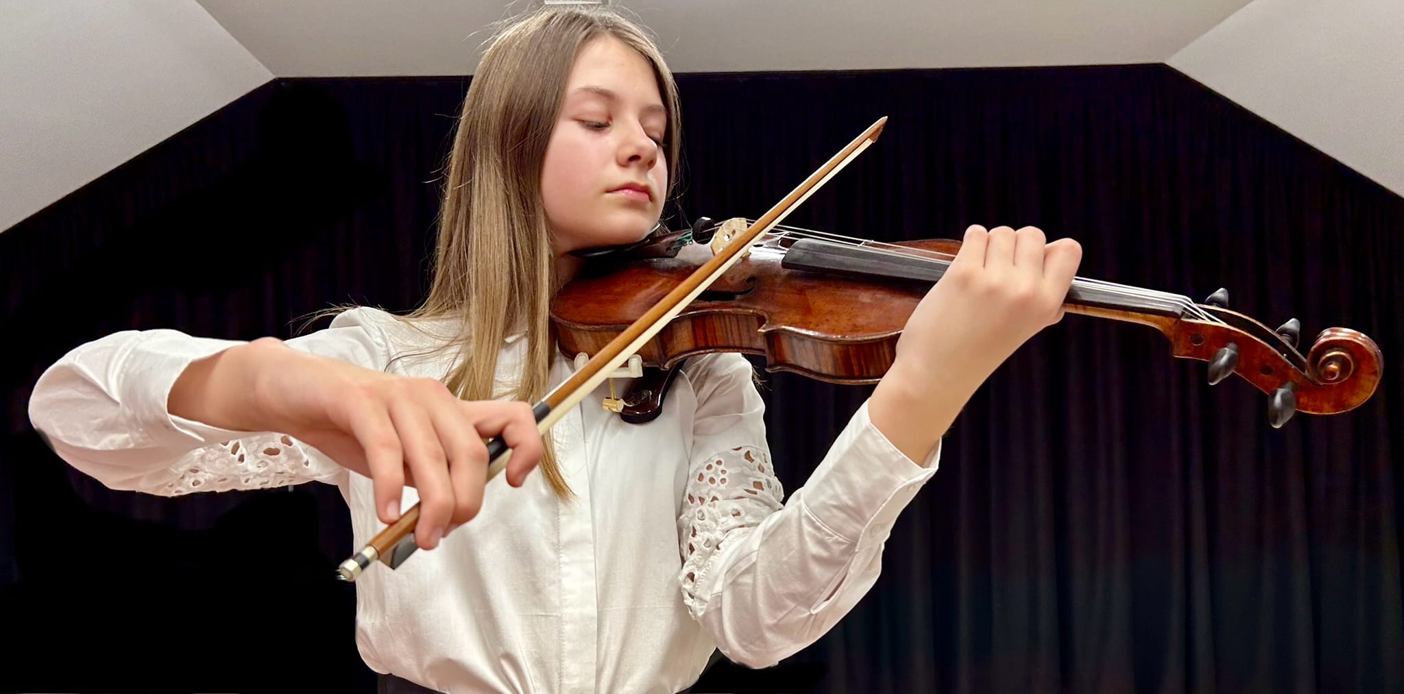 Na zdjęciu widoczna dziewczynka w białej koszuli grająca na skrzypcach na tle czarnej kotary. 