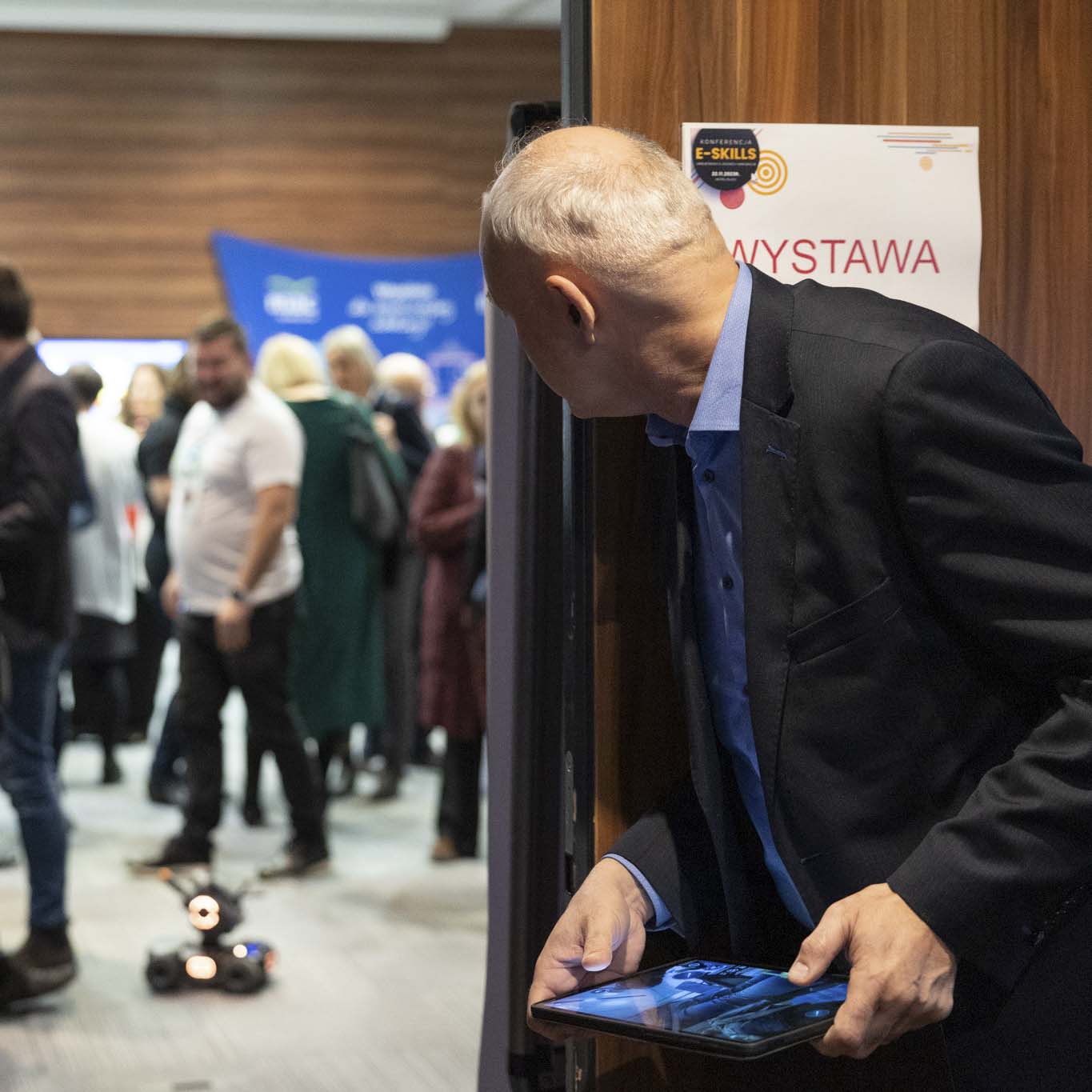 Mężczyzna stoi z tabletem na którym zaprogramował trasę dla robota, śledzi robota wzrokiem. Robot jest na dalszym planie, na sali gdzie jest dużo osób.