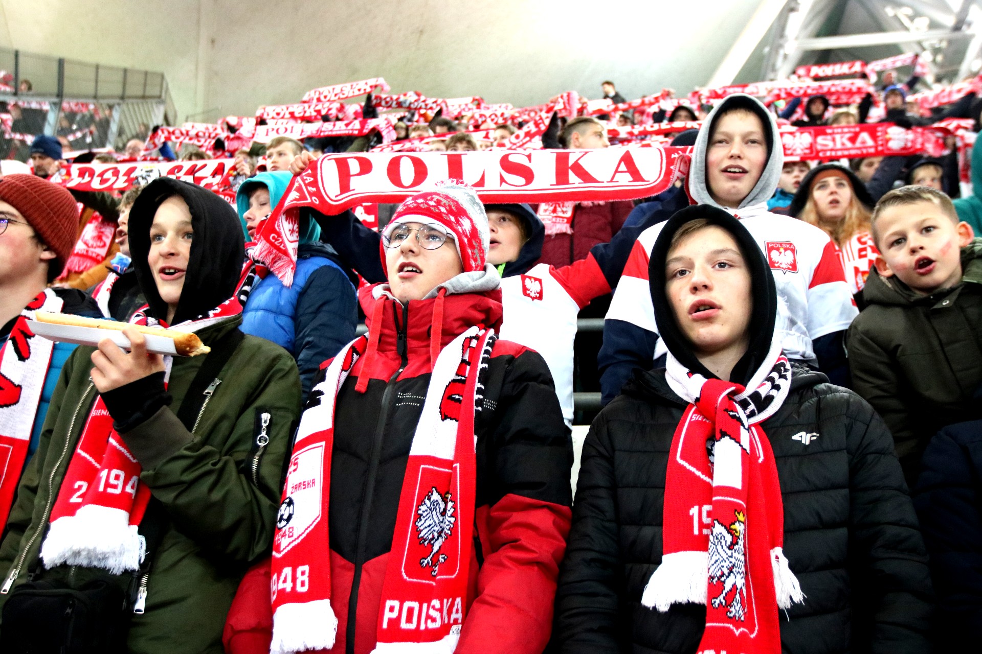 Trybuna stadionu wypełniona dziećmi w biało-czerwonych czapkach i szalikach, trzymają szaliki z napisem Polska.