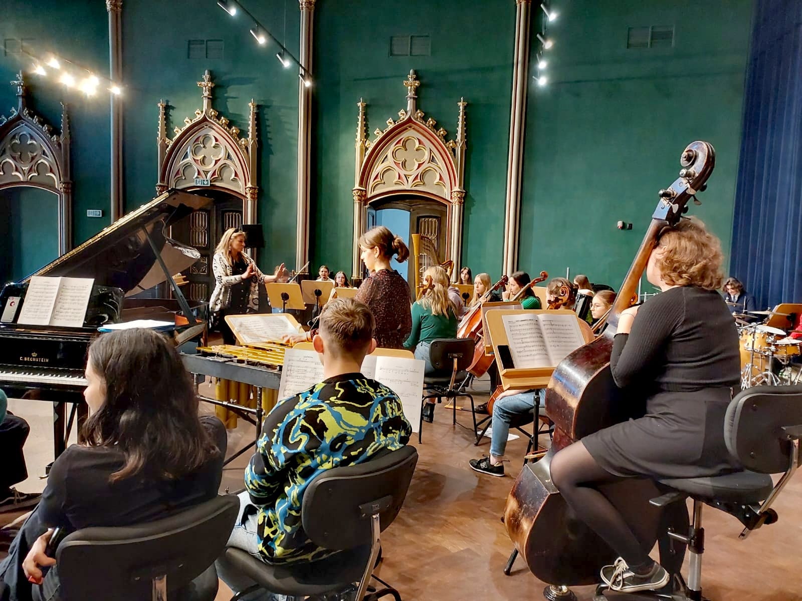 Zdjęcie orkiestry symfonicznej, młodzieżowej. Z prawej strony na pierwszym planie dziewczyna z kontrabasem siedząca na krześle, z lewej otwarty fortepian z rozłożonymi nutami na pulpicie. Za fortepianem pani dyrygent, przed nią dziewczyna grająca na wibrafonie. W oddali wiolonczele skrzypce i harfa. Wszystko to dzieje się w sali koncertowej OSM w Bytomiu, ściany w kolorze zielonym(butelkowym) z pięknie zdobionymi odrzwiami.