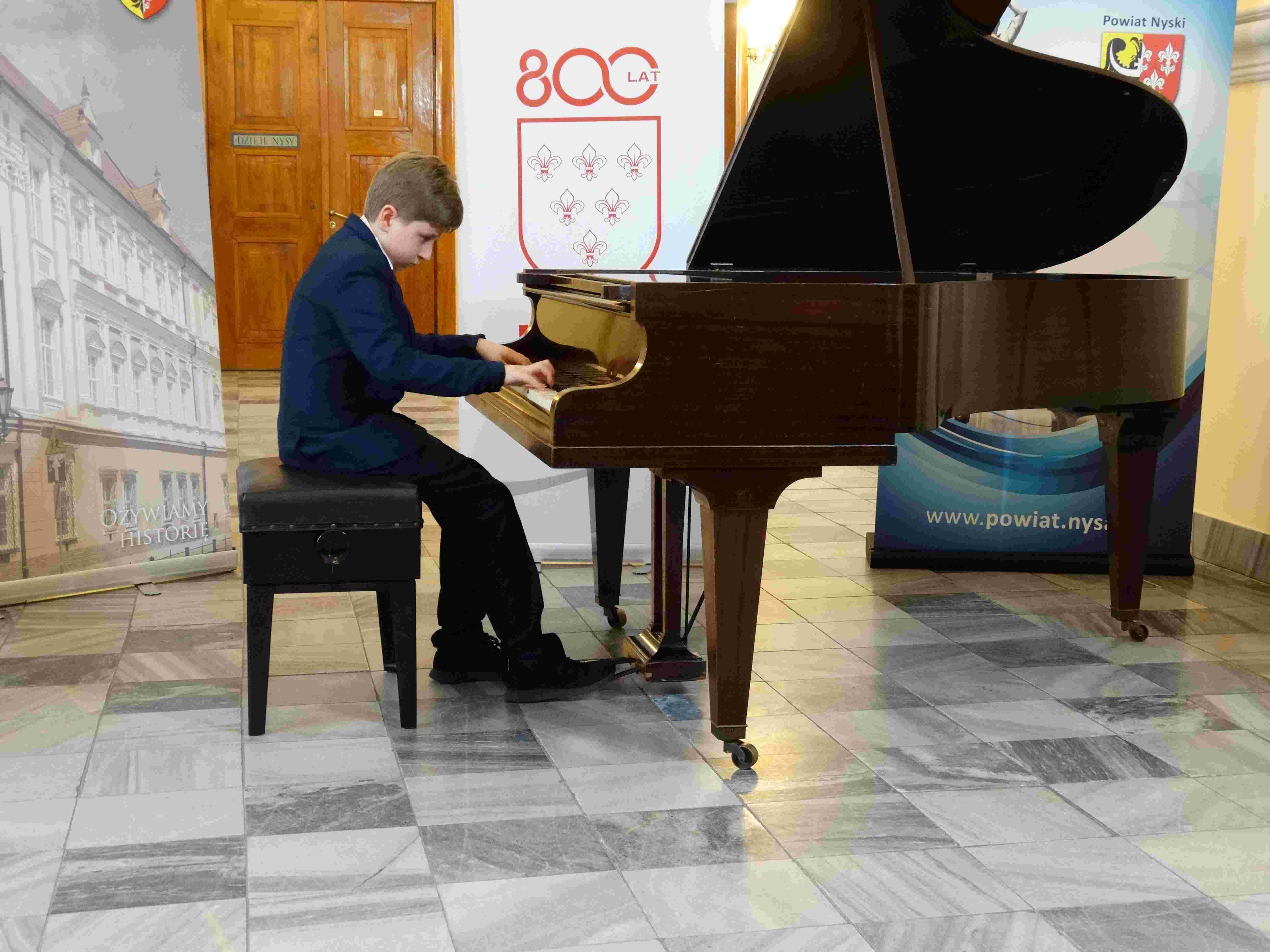 Zdjęcie. Na fortepianie w Muzeum Powiatowym w Nysie gra młody pianista. W tle baner 800-lecia Nysy.