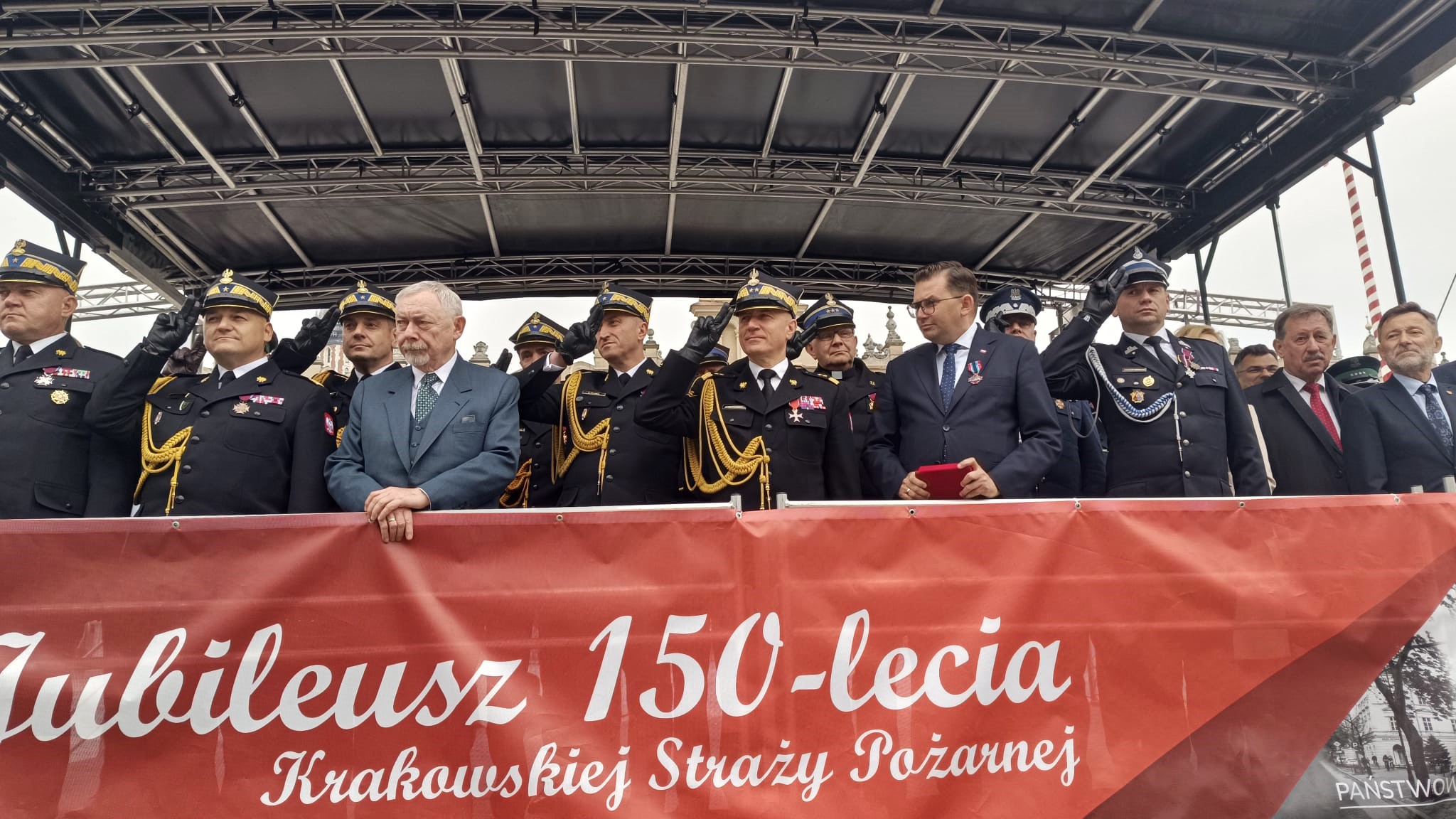 Na scenie stoją strażacy i osoby cywilne, strażacy salutują,. na dole napis na czerwonym tle Jubileusz 150-lecia Krakowskiej Straży Pożarnej